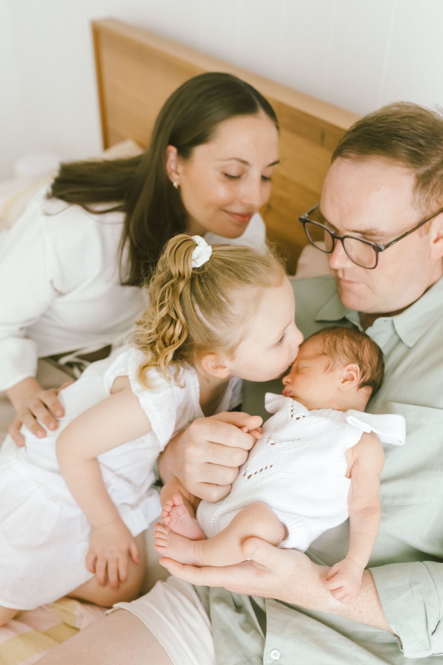 Family with newborn baby, toddler girl kissing infant held by father wearing glasses, mother smiling behind them on bed.