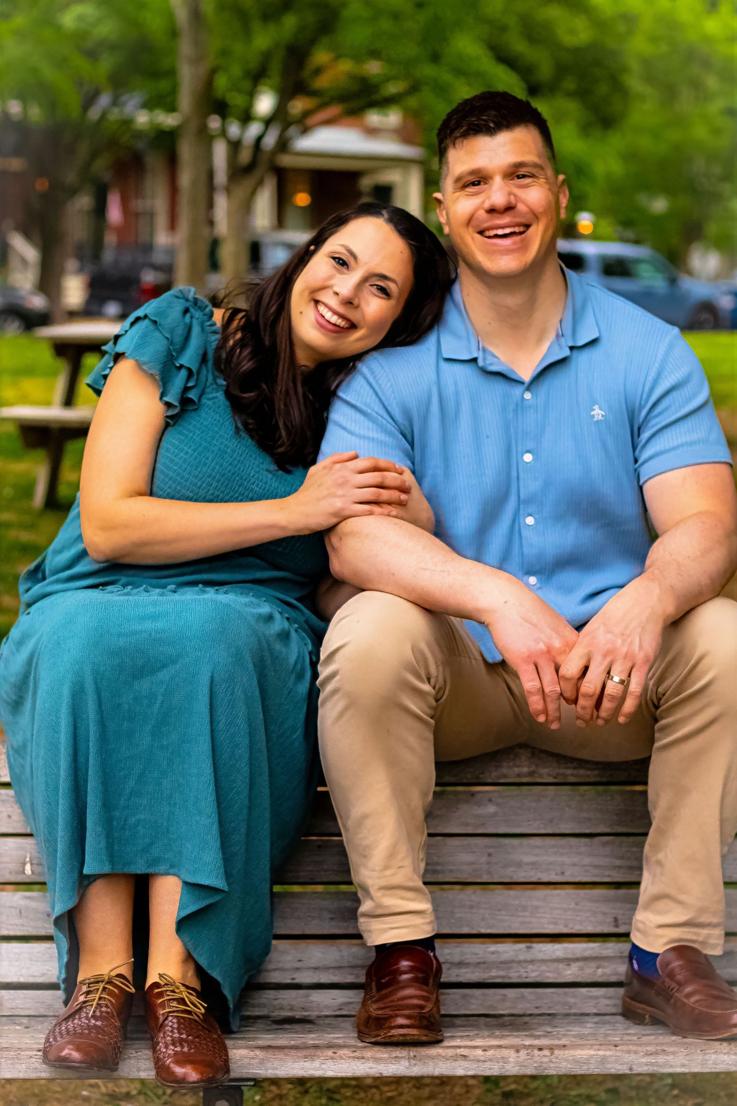 A smiling couple sits closely on a park bench, woman in teal dress leaning on man in blue polo shirt.
