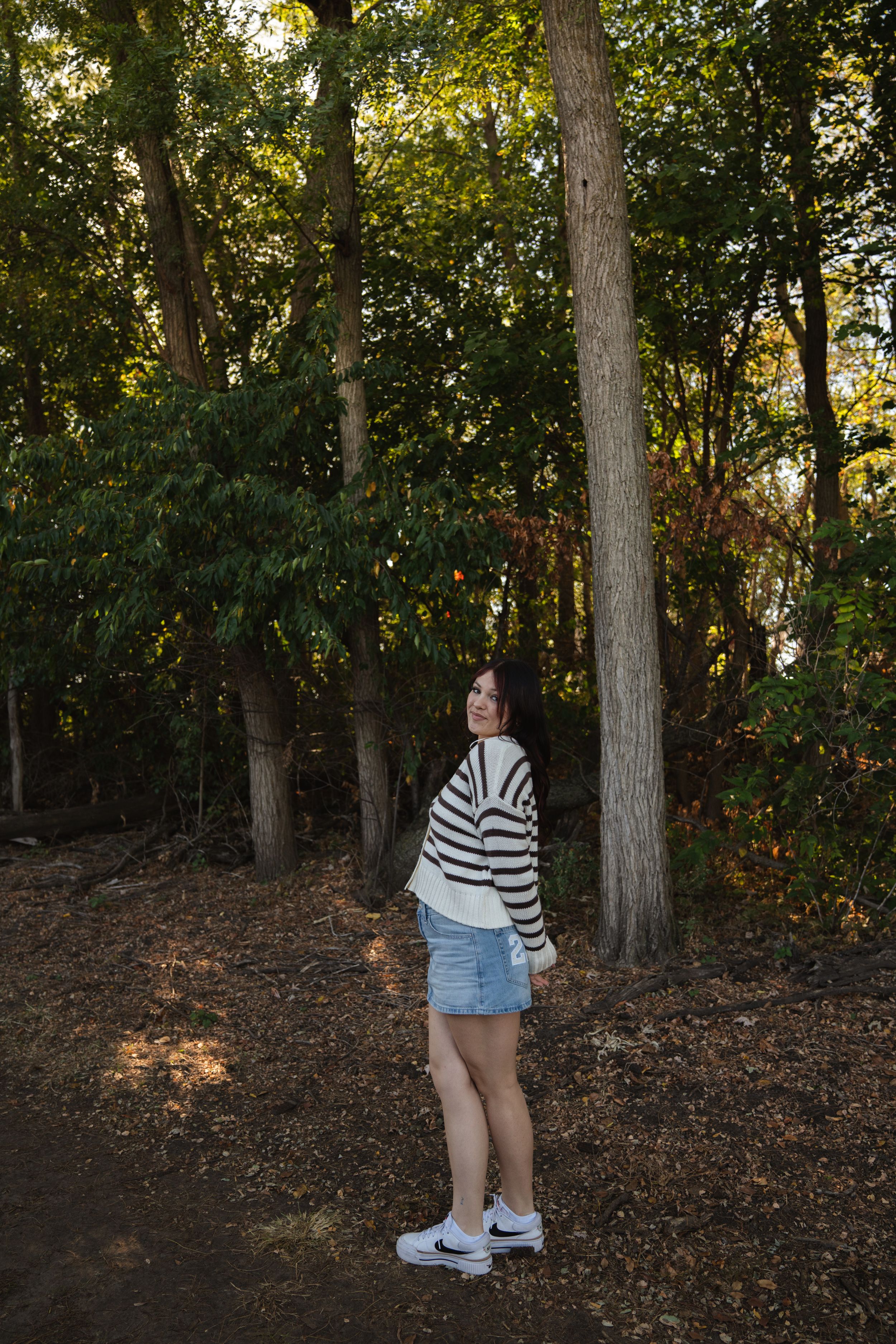Young woman in striped sweater and denim shorts standing on a forest path surrounded by tall trees in autumn.