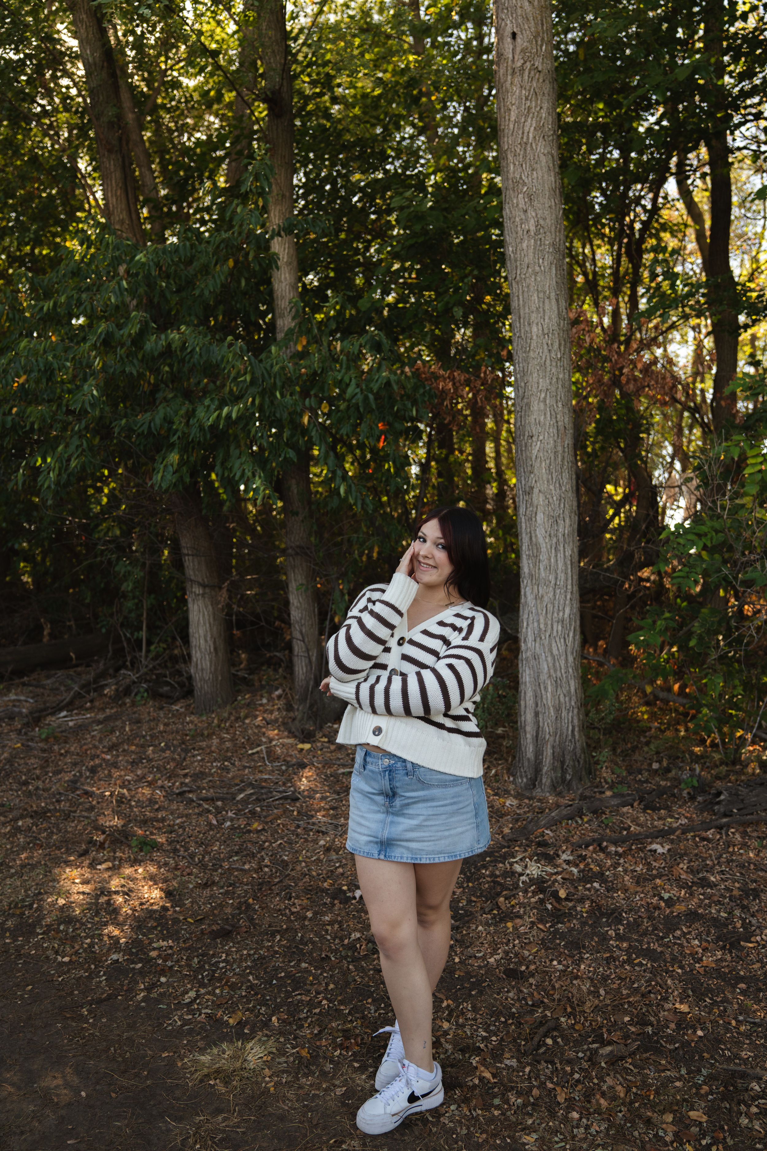 Young woman in striped cardigan and denim skirt standing among trees in autumn forest.
