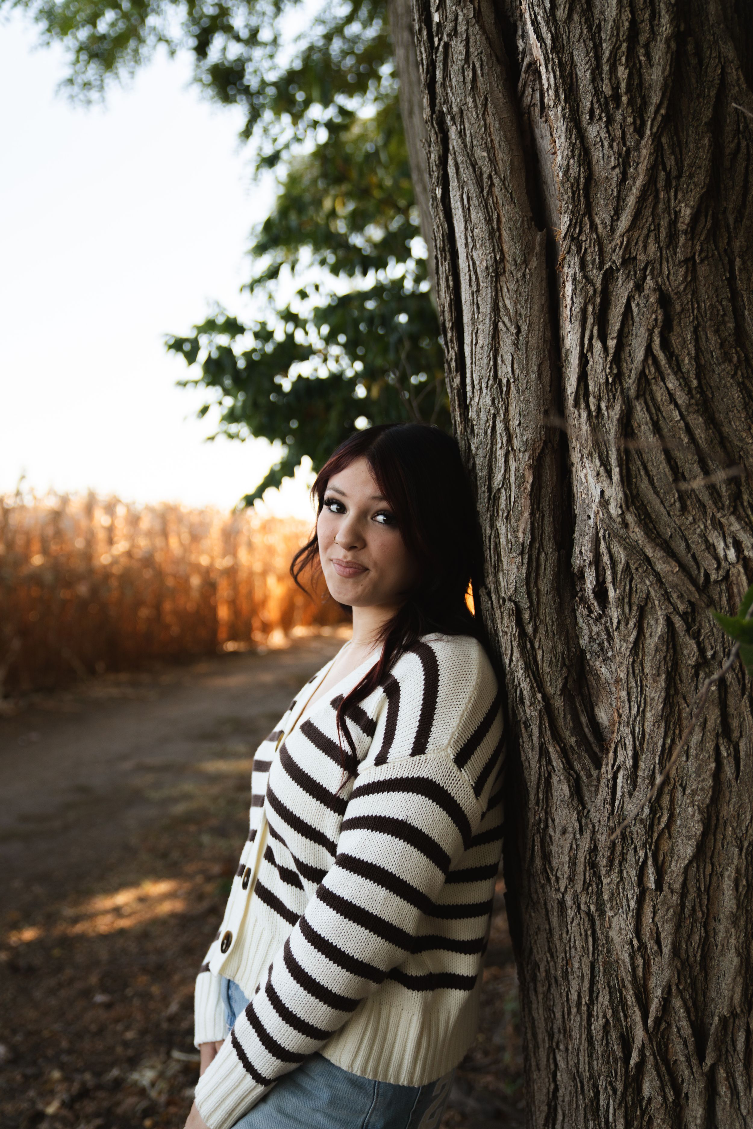 Young woman in striped sweater leaning against a tree trunk outdoors in autumn setting.