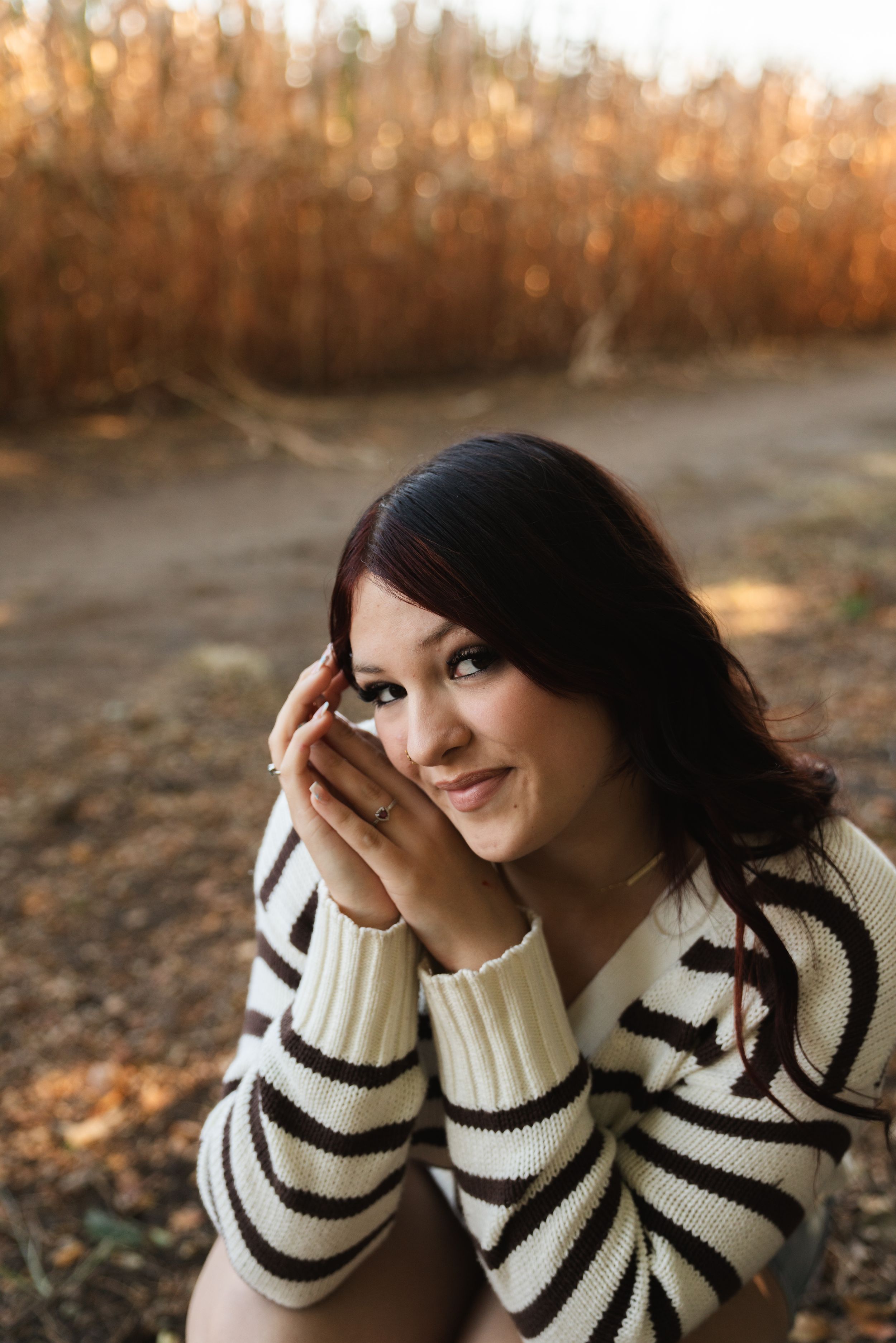 Young woman in striped sweater smiling warmly outdoors near autumn cornfield in golden light.