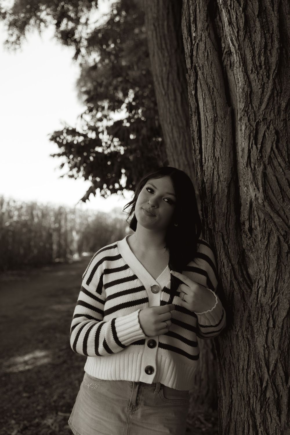 Black and white photo of a young woman leaning against a large tree trunk in a park, wearing a striped cardigan.