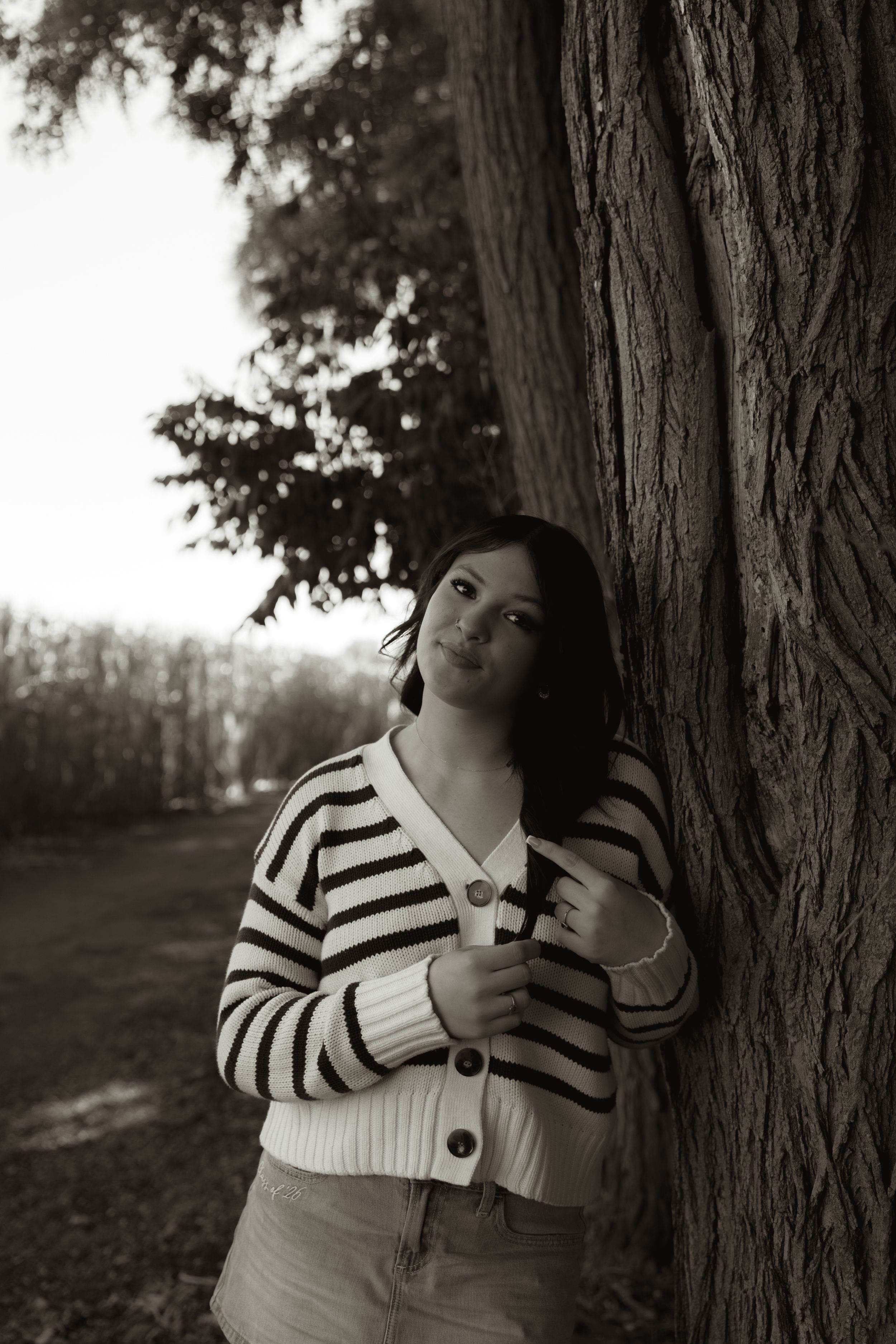 Black and white photo of a young woman leaning against a large tree trunk in a park, wearing a striped cardigan.