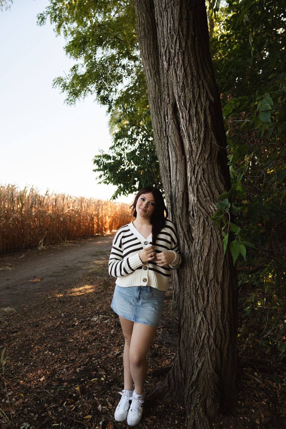 Woman in striped cardigan and denim skirt leaning against tree near cornfield at sunset.