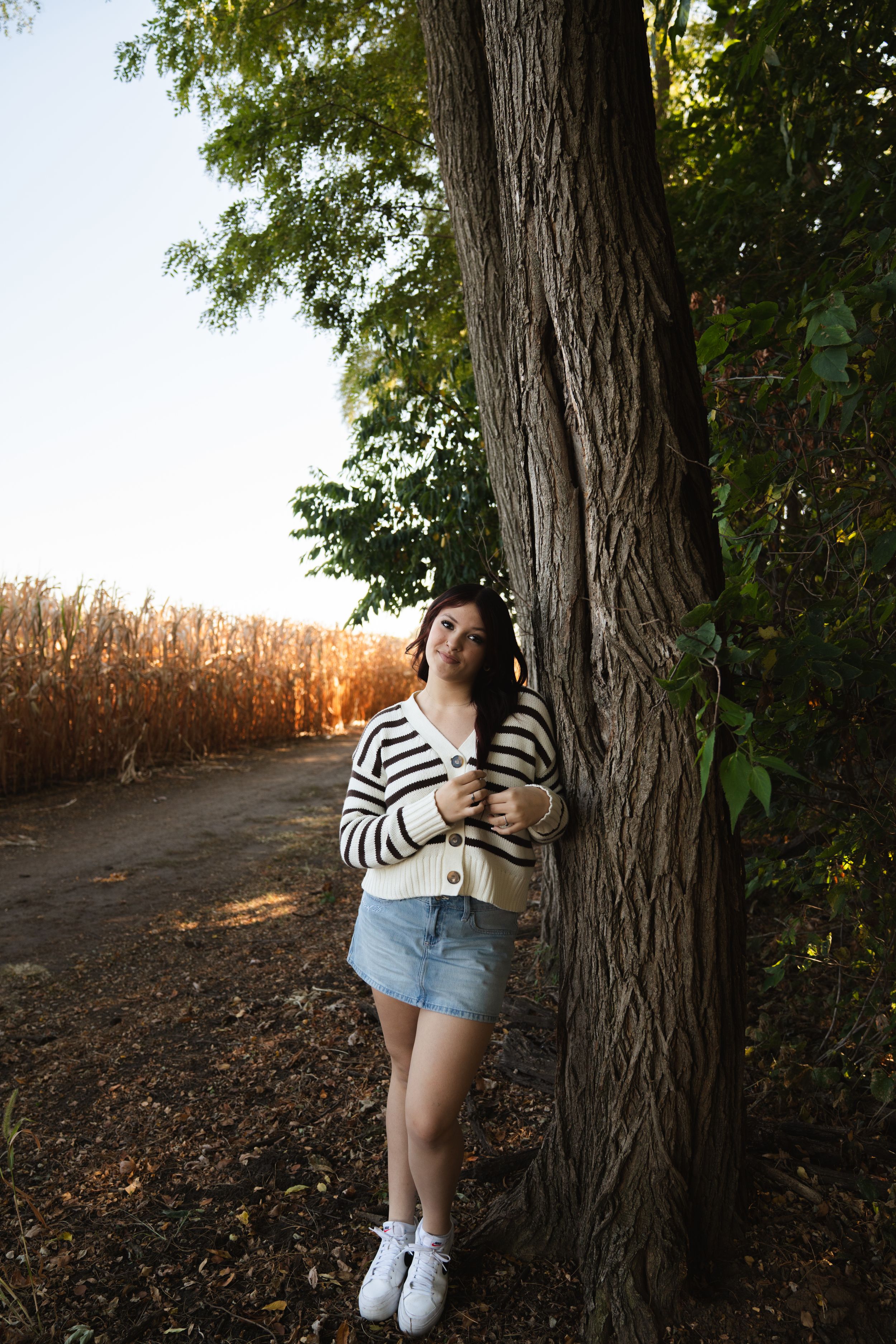 Woman in striped cardigan and denim skirt leaning against tree near cornfield at sunset.