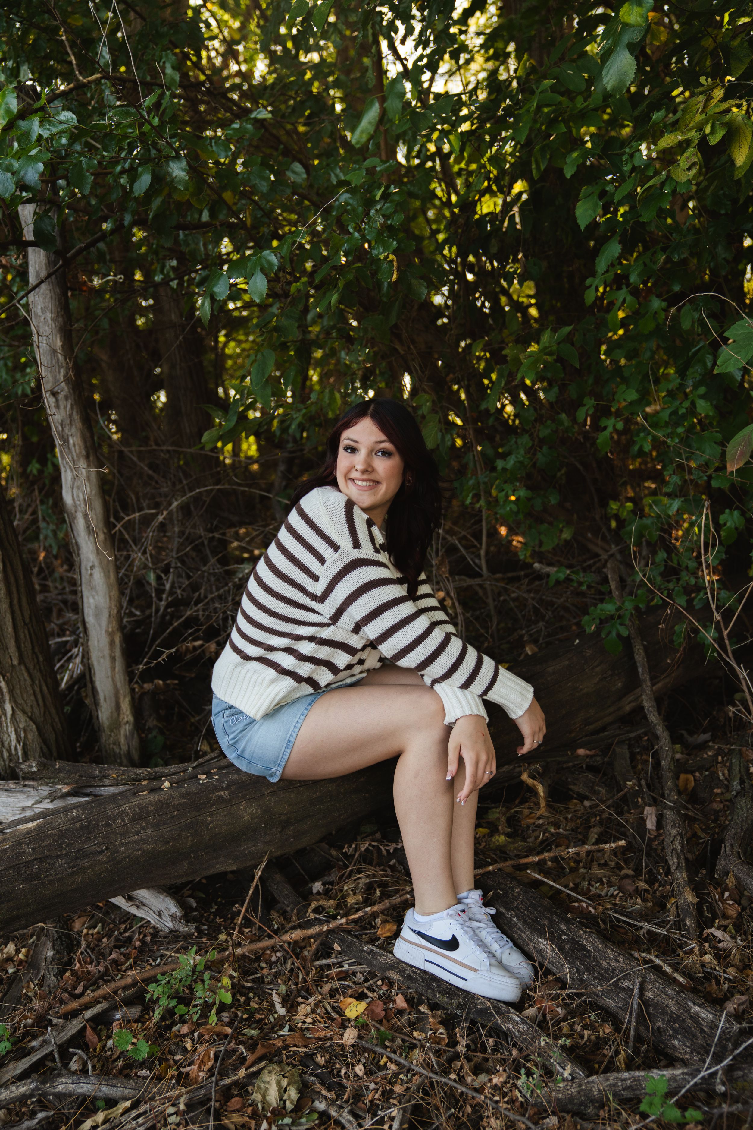 Young woman in striped sweater and denim shorts sitting on a fallen log surrounded by lush green trees.