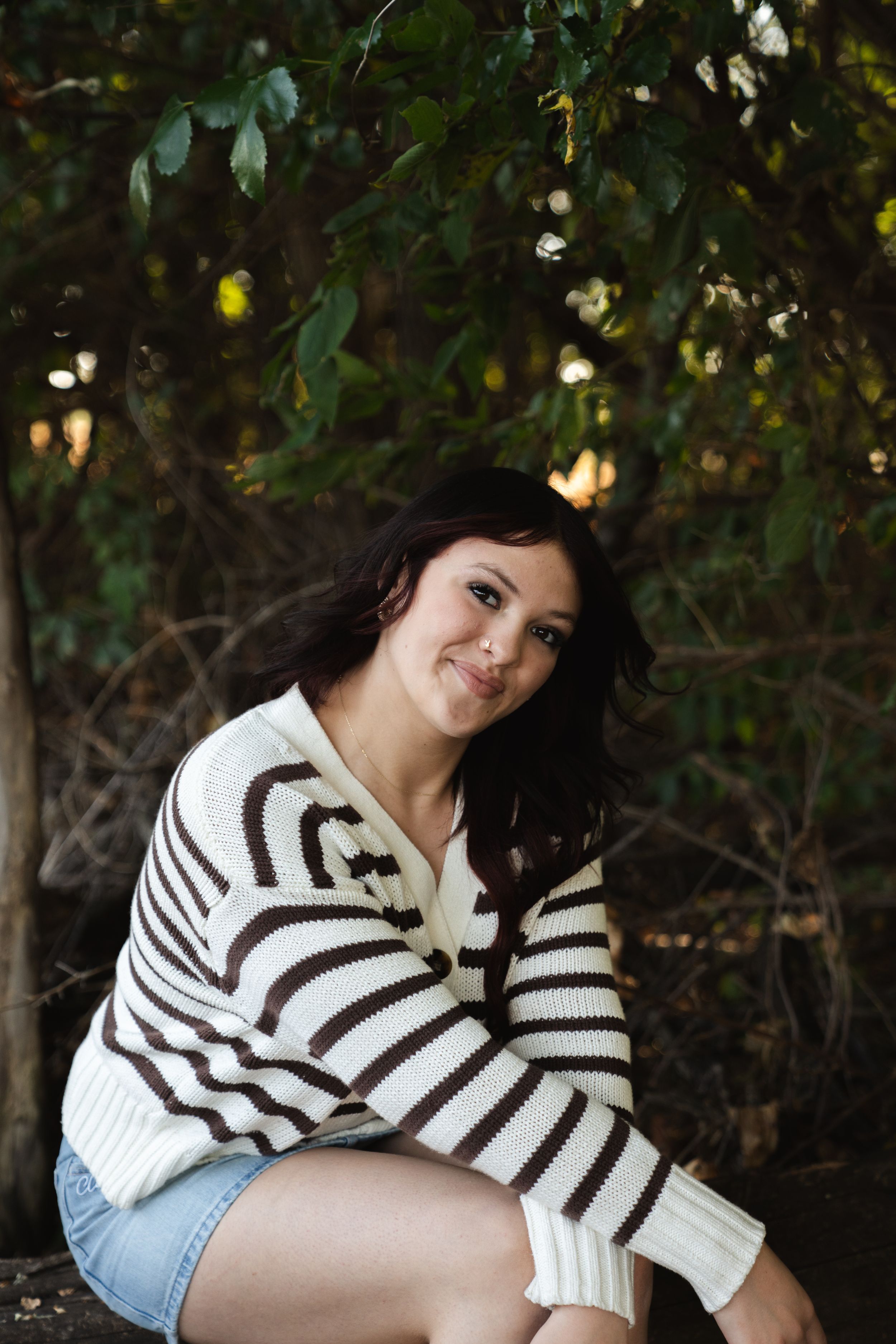 Young woman in striped sweater and denim shorts sitting outdoors near green trees, smiling at camera.