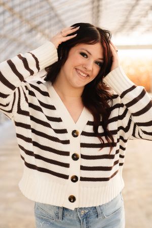 Smiling young woman in striped cream and brown cardigan sweater with jeans, posing outdoors in warm light.