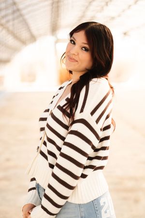 Young woman in striped knit cardigan and jeans smiling over shoulder in warm sunlit outdoor setting.