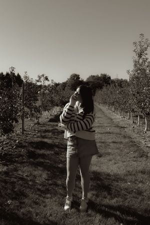 Black and white photo of a woman standing in an orchard pathway, wearing a striped sweater, looking up at the sky.