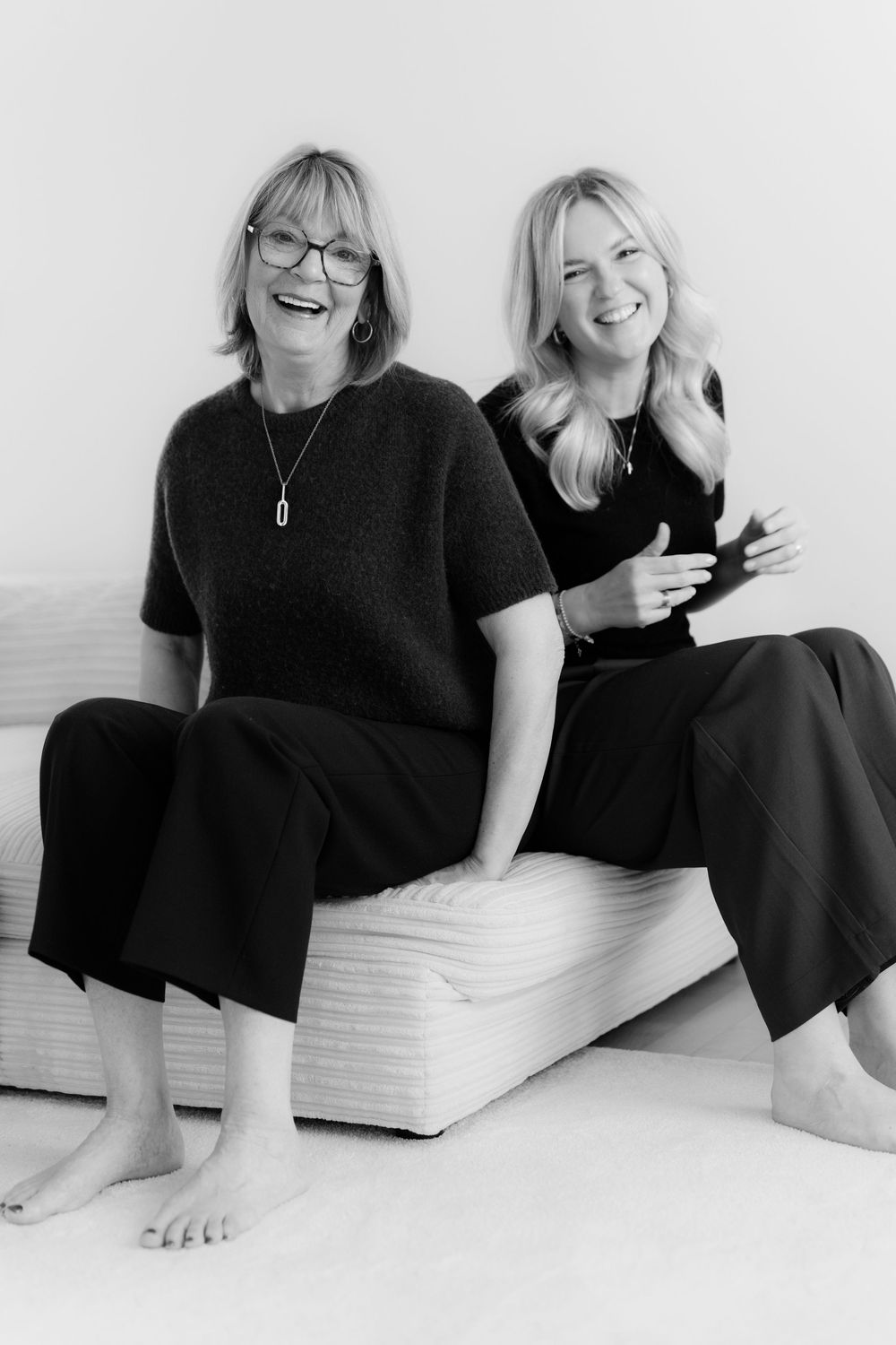 Two women in black outfits sitting barefoot on a white couch, laughing and smiling in a black and white photo.