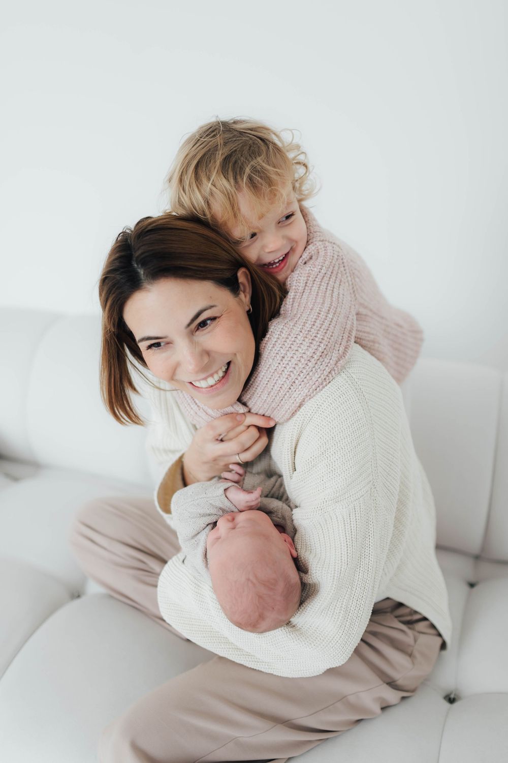 Happy mother holding newborn baby while toddler daughter hugs her from behind on white sofa.