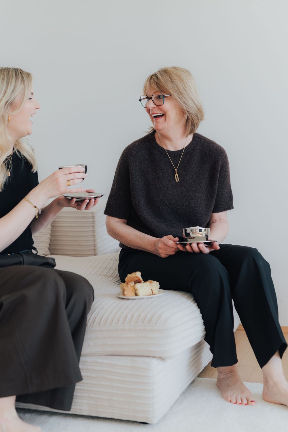 Two women laughing and sharing tea and snacks while seated on white cushioned ottomans in a bright, minimal space.
