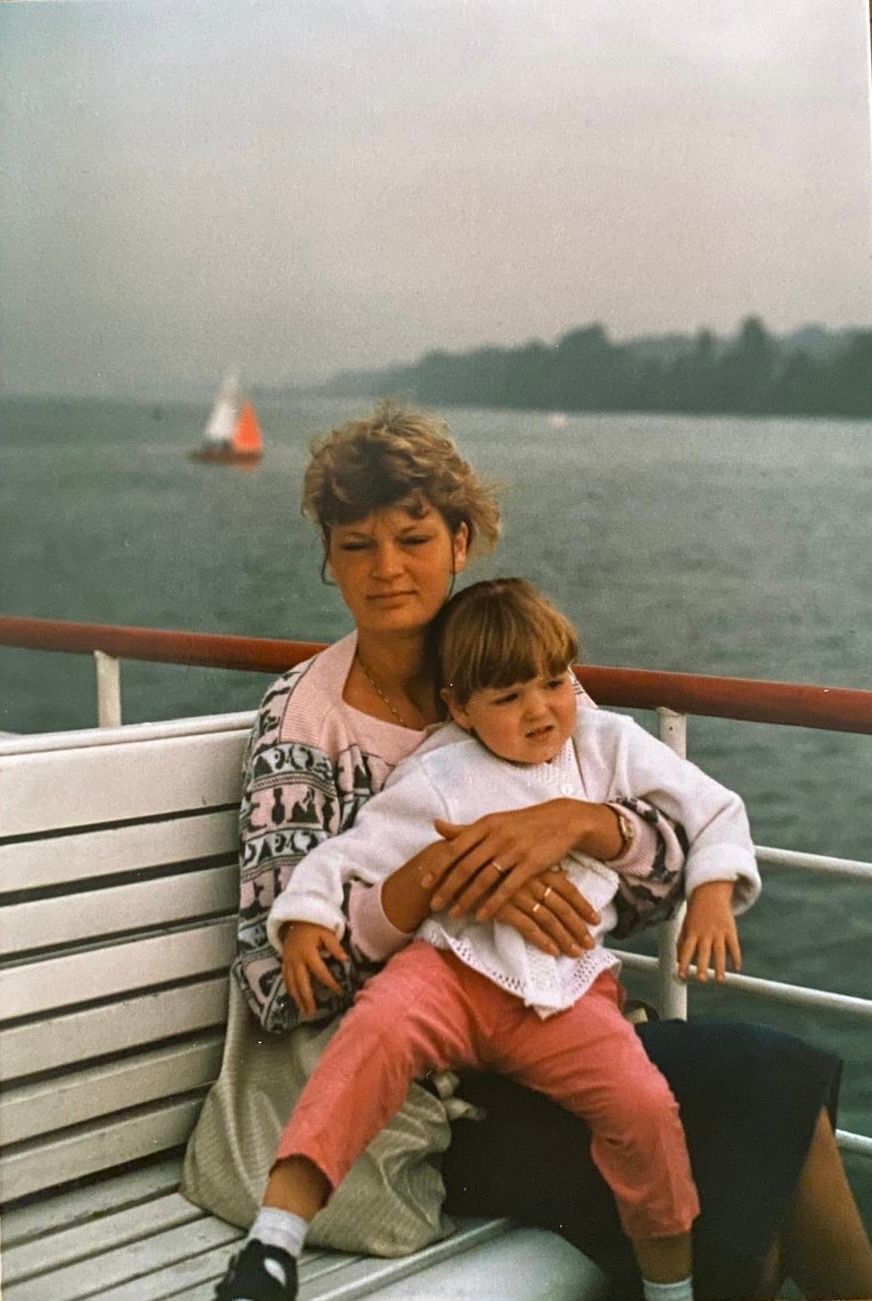 A woman and young child sit together on a bench aboard a boat, with a sailboat visible on the water behind them.