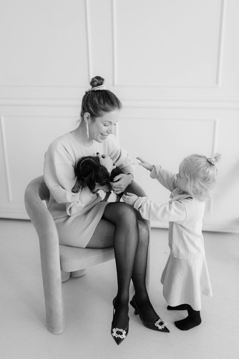 Black and white photo of a woman sitting in a chair holding a small dog while a toddler reaches out to pet it.