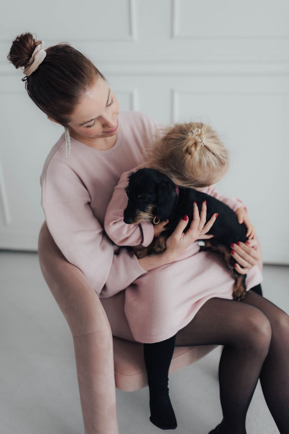 Woman in pink dress sits on chair hugging young blonde child holding a black puppy in bright white room.