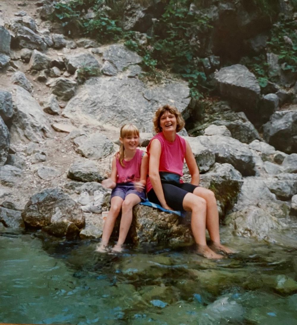 Two girls in pink tops sitting on rocks with feet in a clear mountain stream surrounded by rocky terrain.