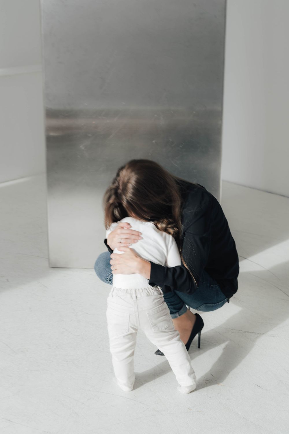 A woman in jeans and heels kneels on a white floor, tenderly embracing a small toddler near a silver wall.