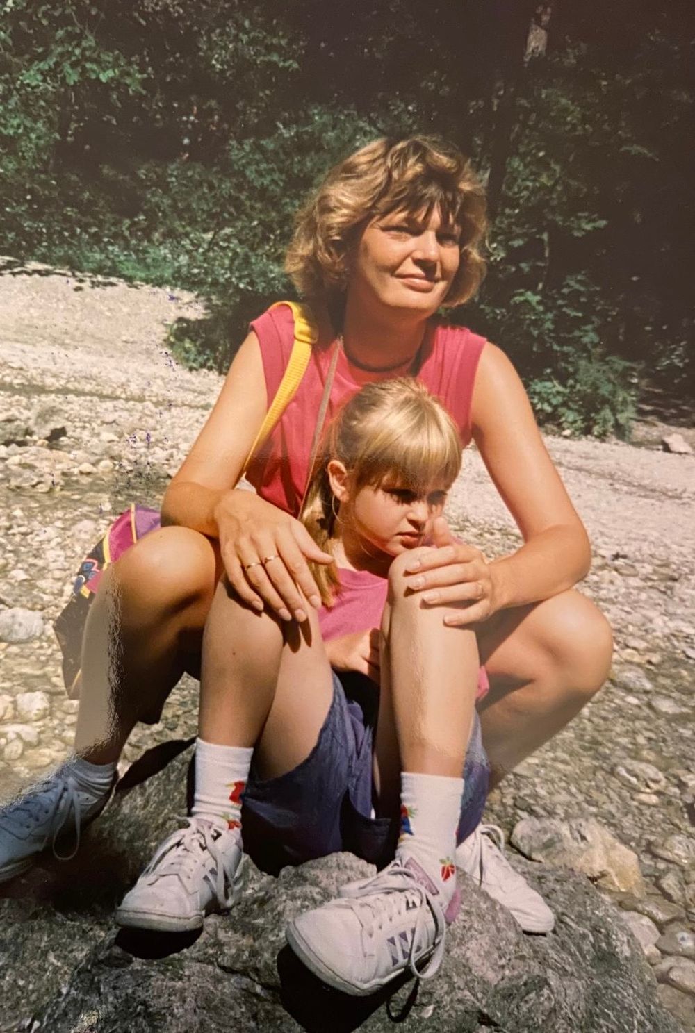 A woman and young girl sit together on rocks near a stream, both wearing pink shirts and white sneakers.