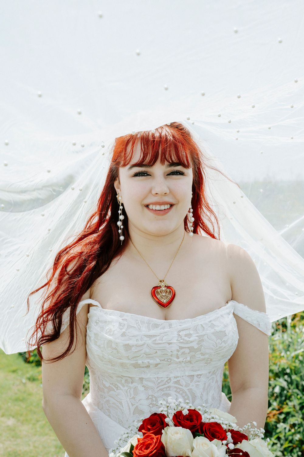 Smiling bride with red hair wearing a white lace off-shoulder gown, heart necklace, and holding a red rose bouquet.