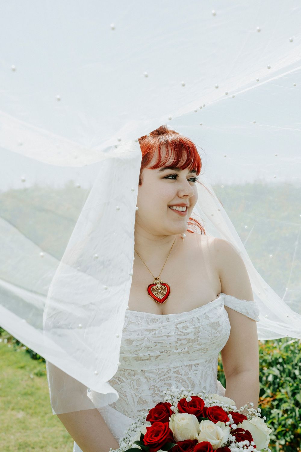 Smiling bride in white off-shoulder lace gown with red heart necklace, holding red roses bouquet under flowing veil outdoors.