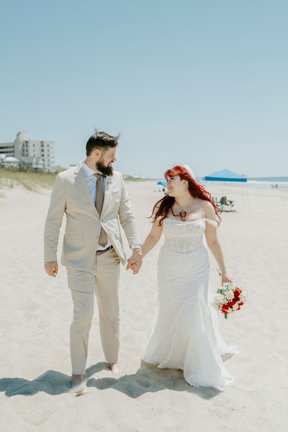 Bride and groom walk hand-in-hand on a sunny beach, she in a white gown with red bouquet, he in a tan suit.
