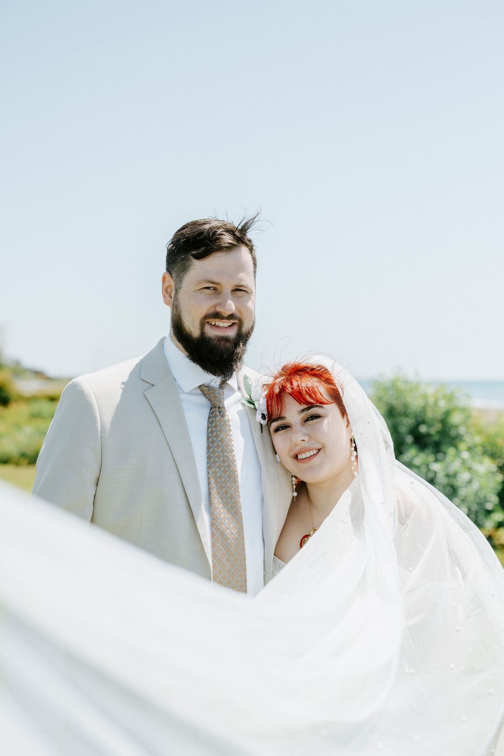 Smiling bride with red hair and groom in gray suit pose outdoors on their wedding day with veil flowing in the breeze.