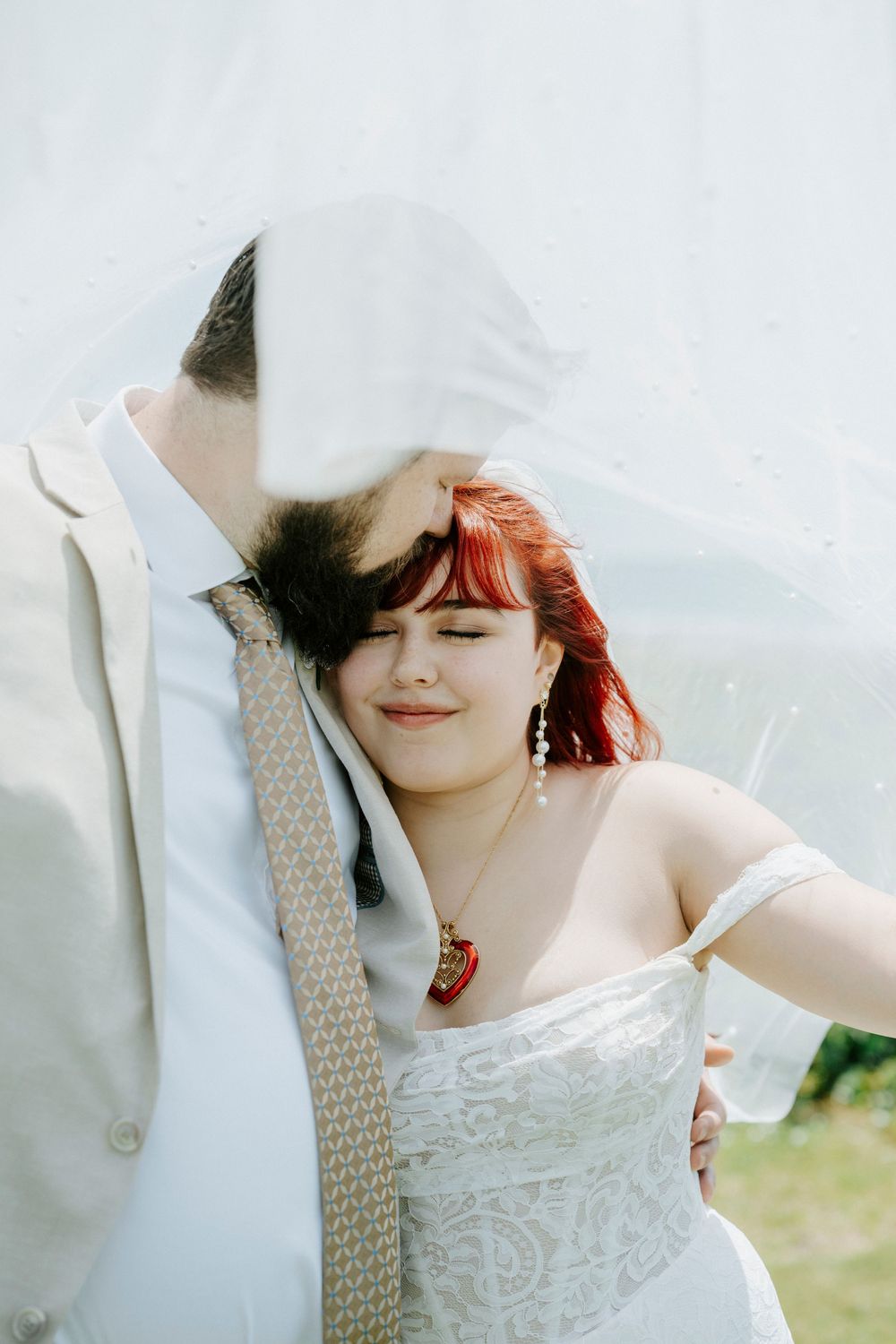 Bride with red hair leans on groom's shoulder under veil, wearing white dress and heart necklace at outdoor wedding.