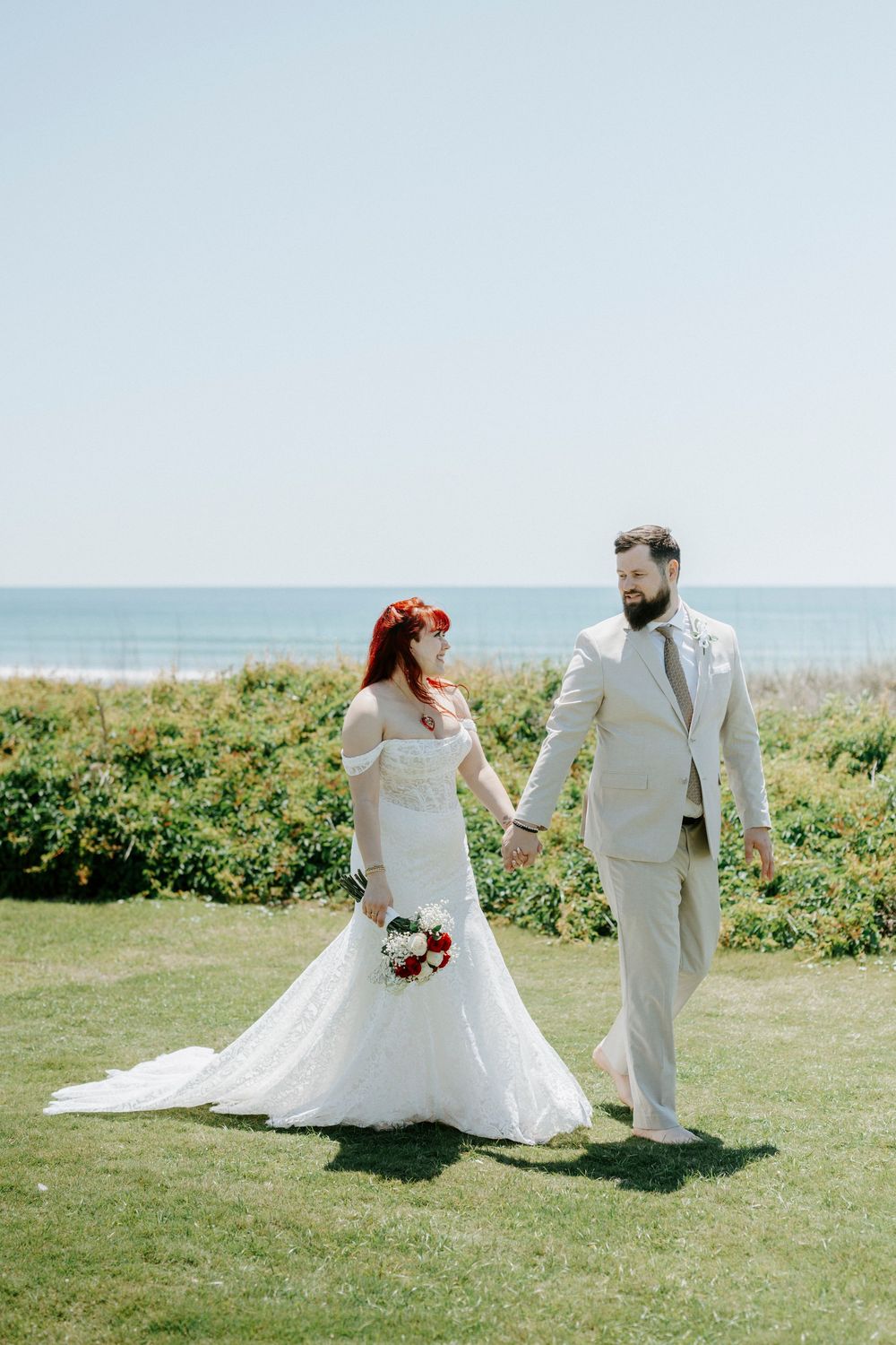 A bride with red hair and groom in tan suit hold hands on green lawn with ocean backdrop on their wedding day.