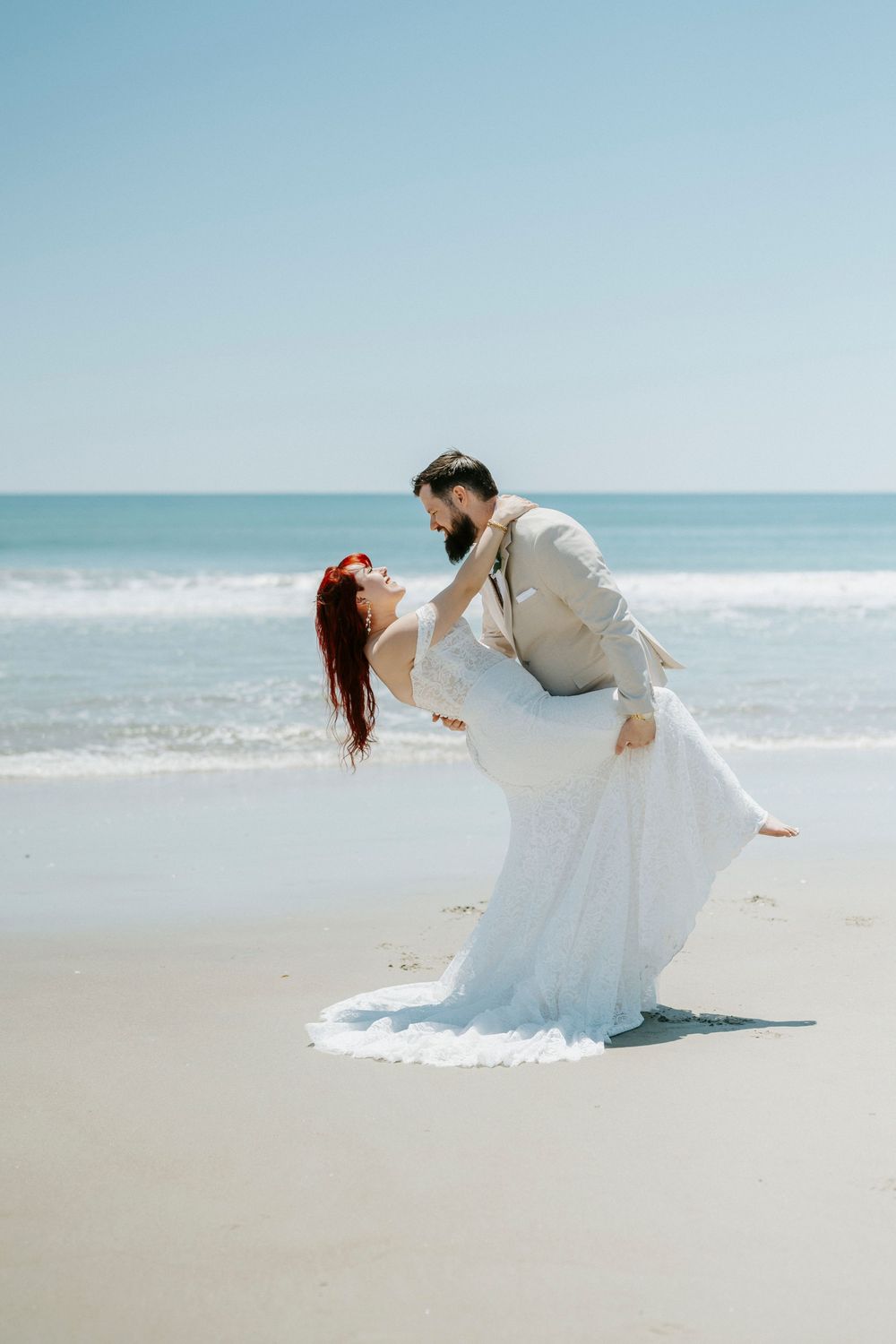 Bride in white gown and groom in tan suit share romantic dip on sunny beach with ocean waves behind them.