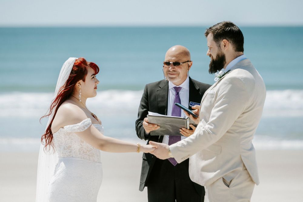 Couple exchanging vows at a beach wedding ceremony with an officiant, bride in white dress, groom in tan suit.