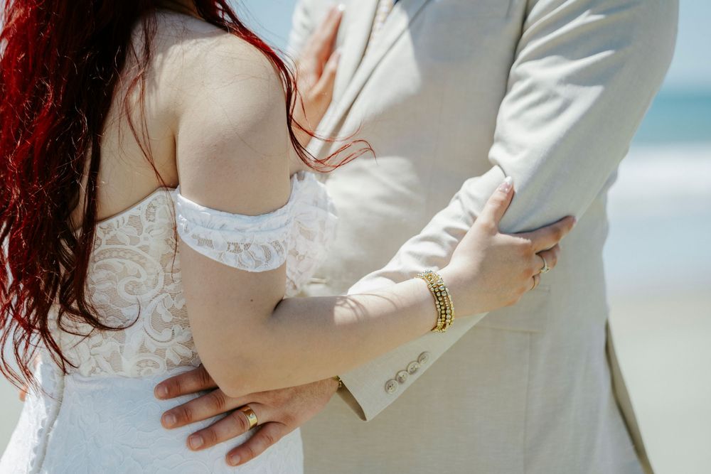 Couple embracing on a beach wedding, bride in white lace dress with gold bracelet, groom in beige suit.