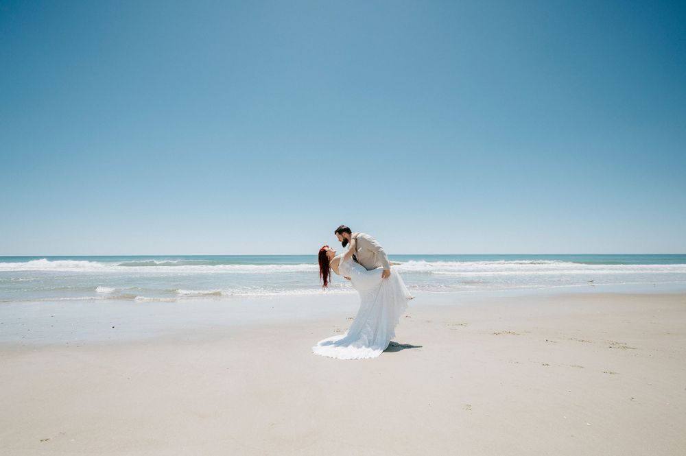 Couple sharing a romantic dip kiss on a sandy beach with ocean waves and clear blue sky on their wedding day.