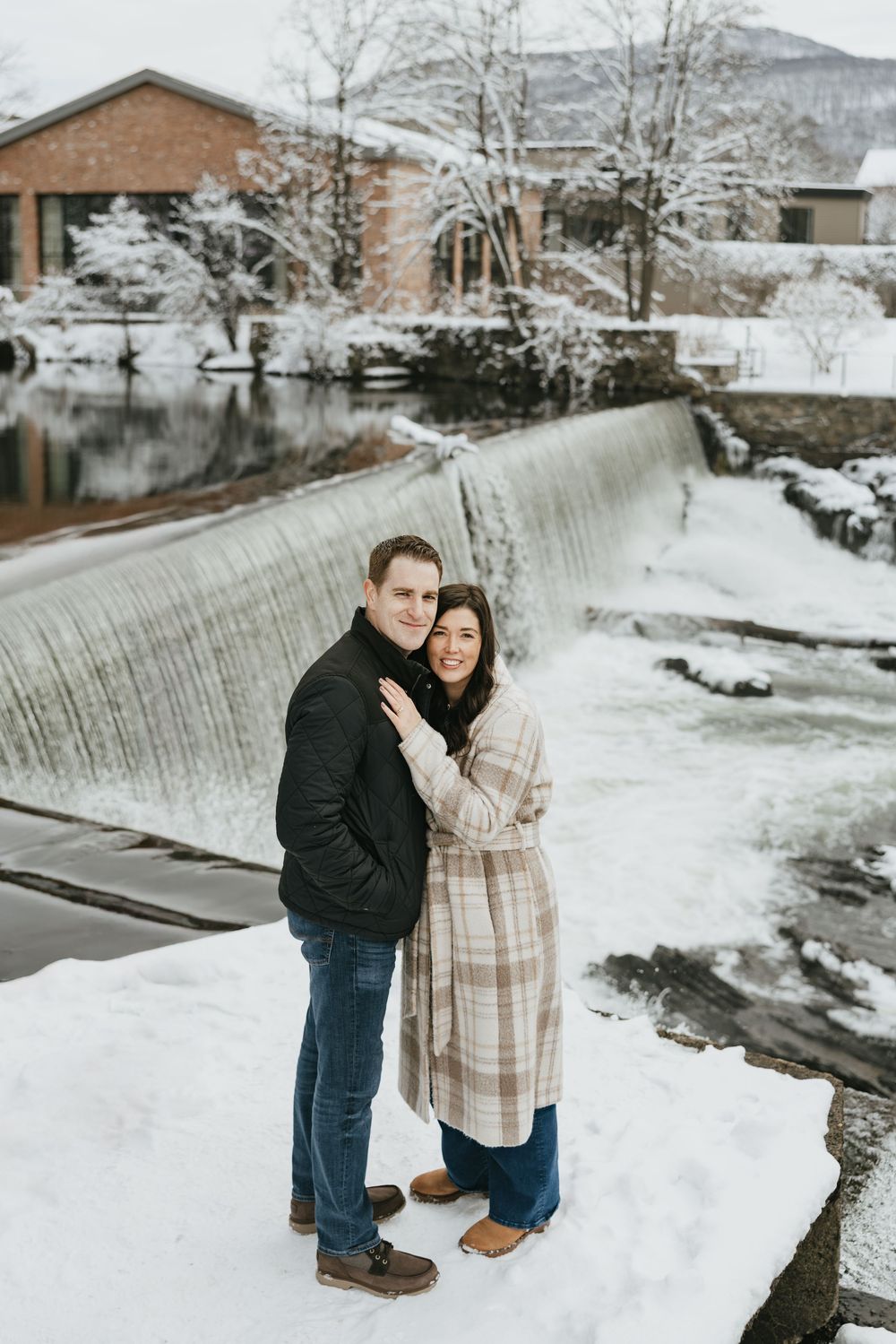 Couple embracing in snow by a waterfall, wearing winter coats, smiling outdoors near a brick building.