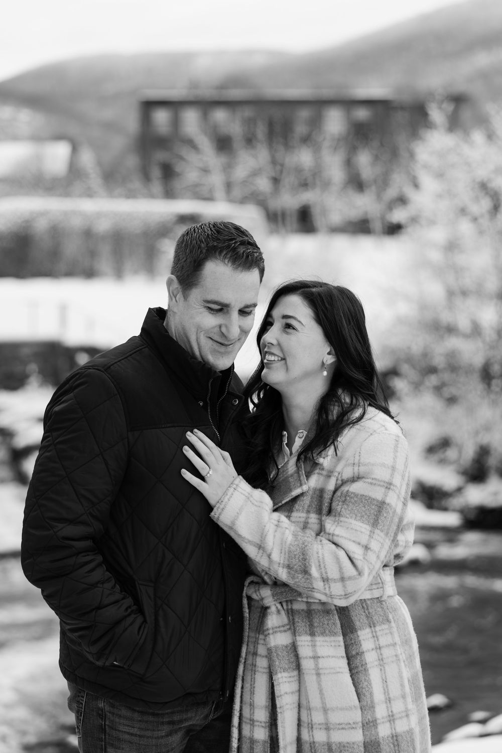 Black and white photo of a smiling couple embracing outdoors in winter, with snowy mountains in the background.