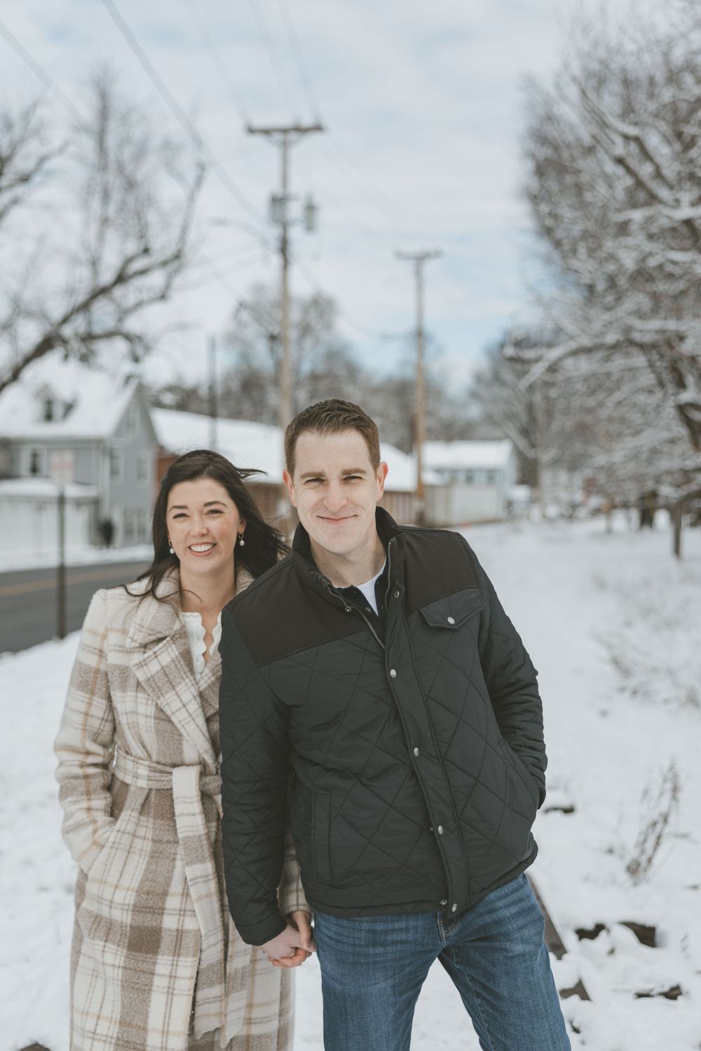 Couple holding hands and smiling in a snowy winter street, woman in plaid coat and man in black quilted jacket.