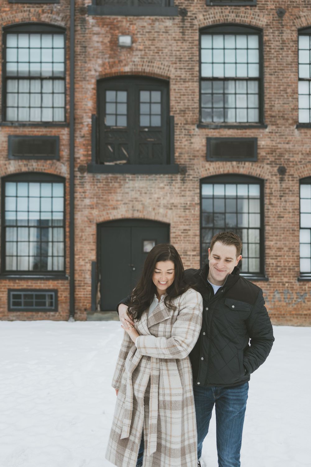 Couple smiling together in winter outside a brick building with snow on the ground, woman in plaid coat.