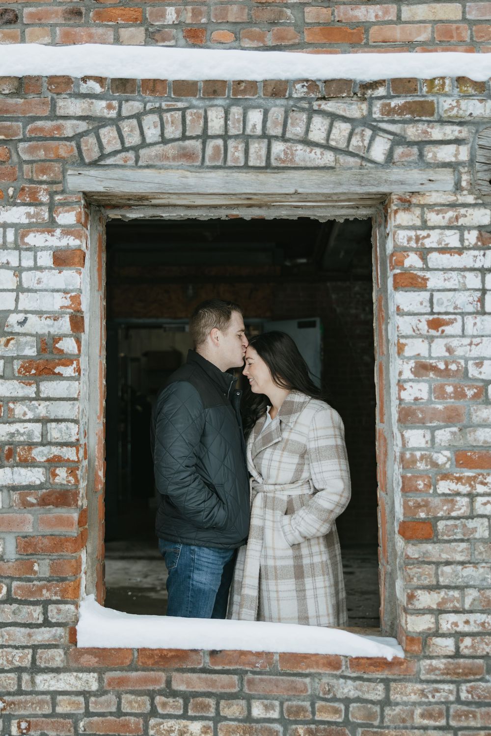 Couple sharing a romantic kiss in a snow-dusted brick window frame of a rustic building in winter.
