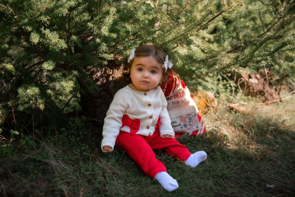 Baby girl in white cardigan and red pants sits on grass under evergreen trees wearing a white bow.