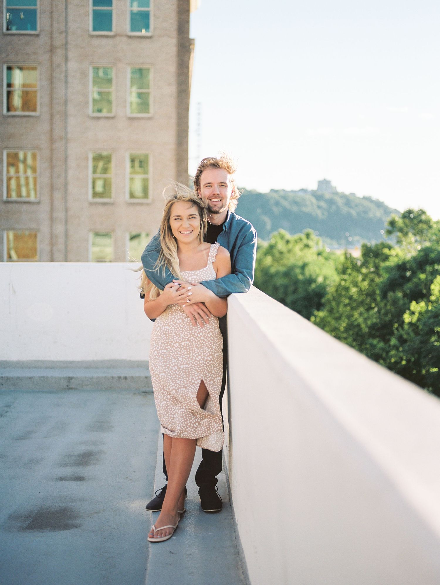 Couple embracing on rooftop with city building and green hills in background on a sunny day.