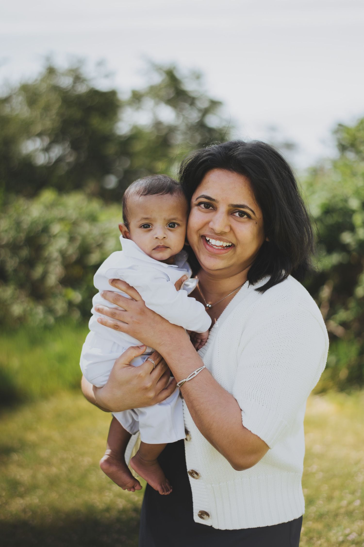 Smiling woman in white cardigan holding a baby boy outdoors with green trees in the background.