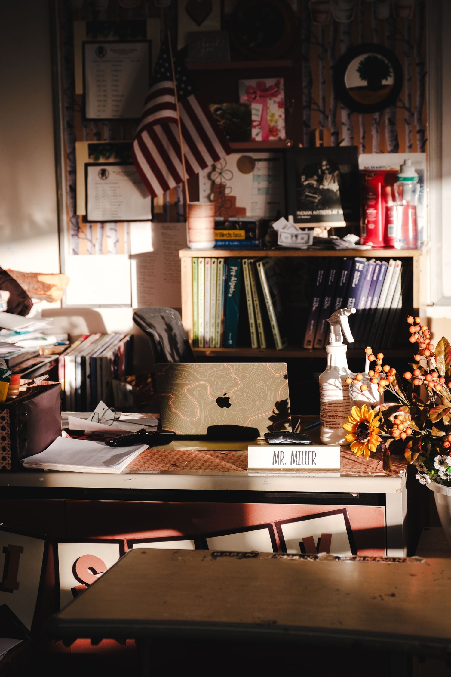 Mr. Miller's cluttered classroom desk with Apple MacBook, sunflowers, bookshelf, and American flag in warm light.