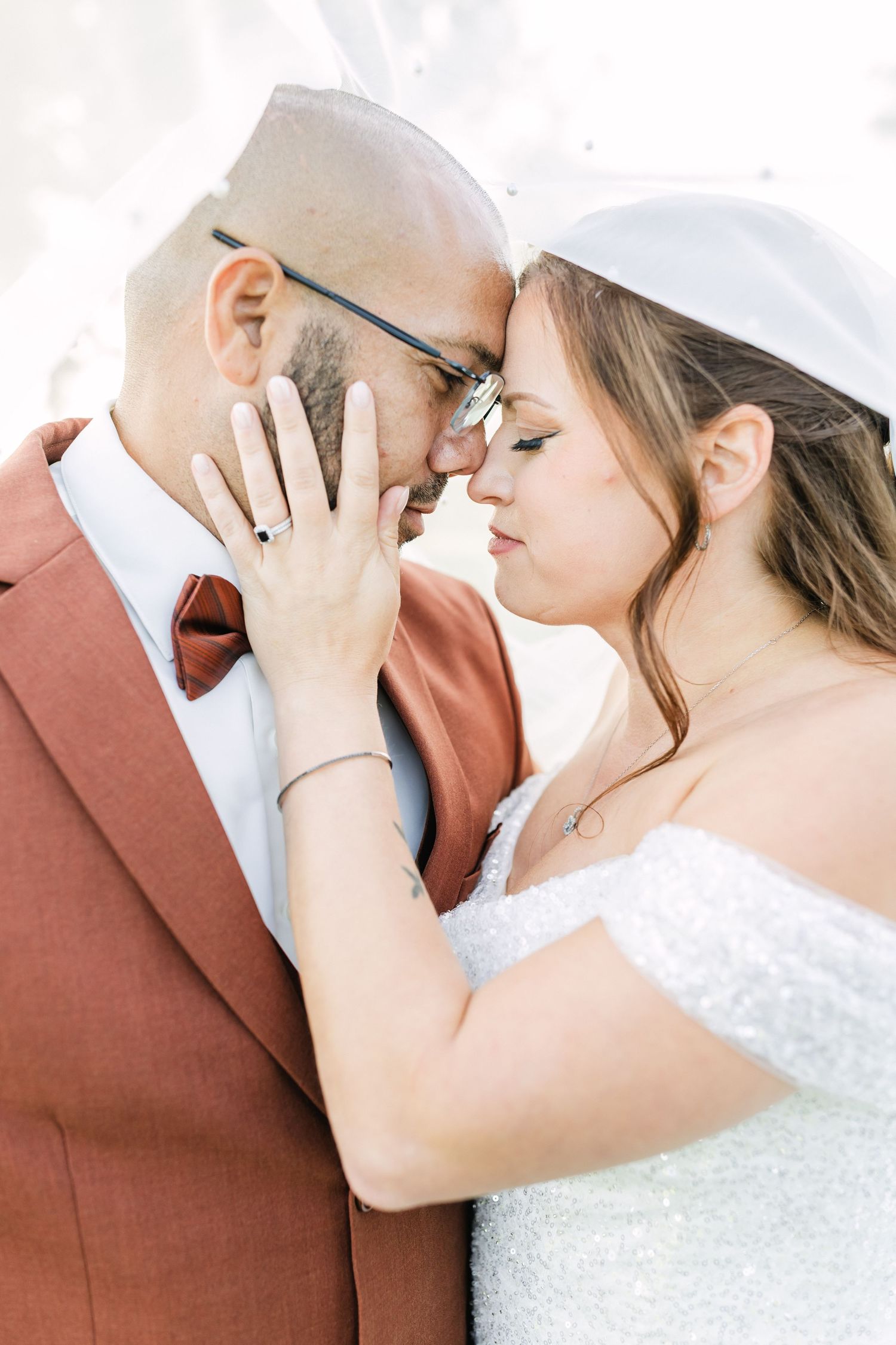 Bride and groom touching foreheads tenderly, groom in rust suit, bride in white lace gown.