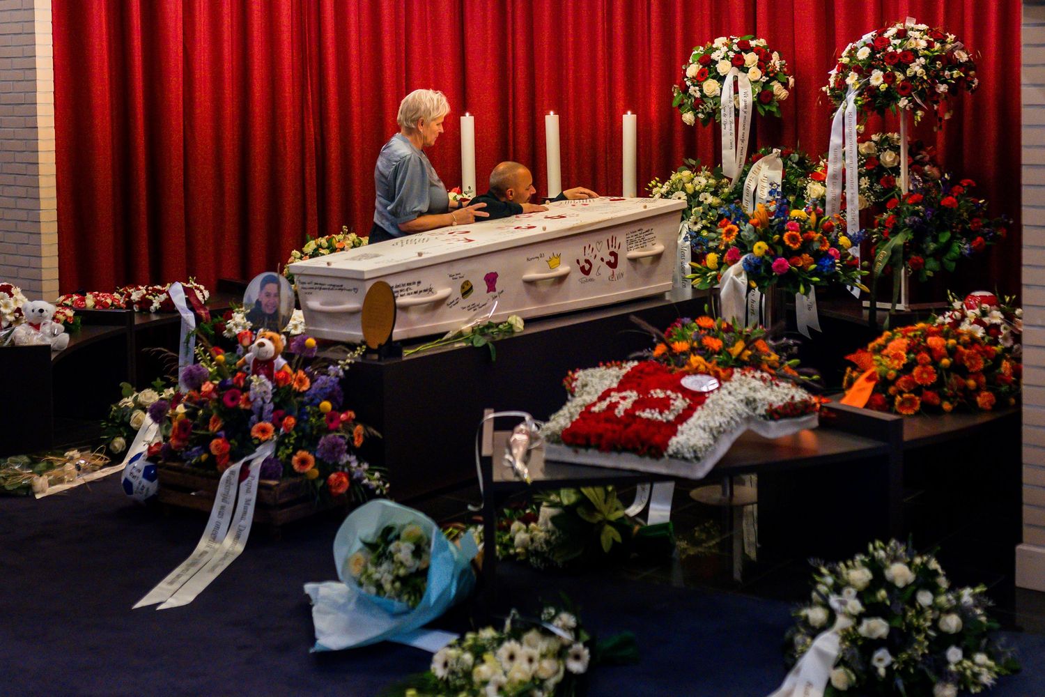 A funeral service with a white casket surrounded by colorful floral arrangements and mourners in a decorated hall.