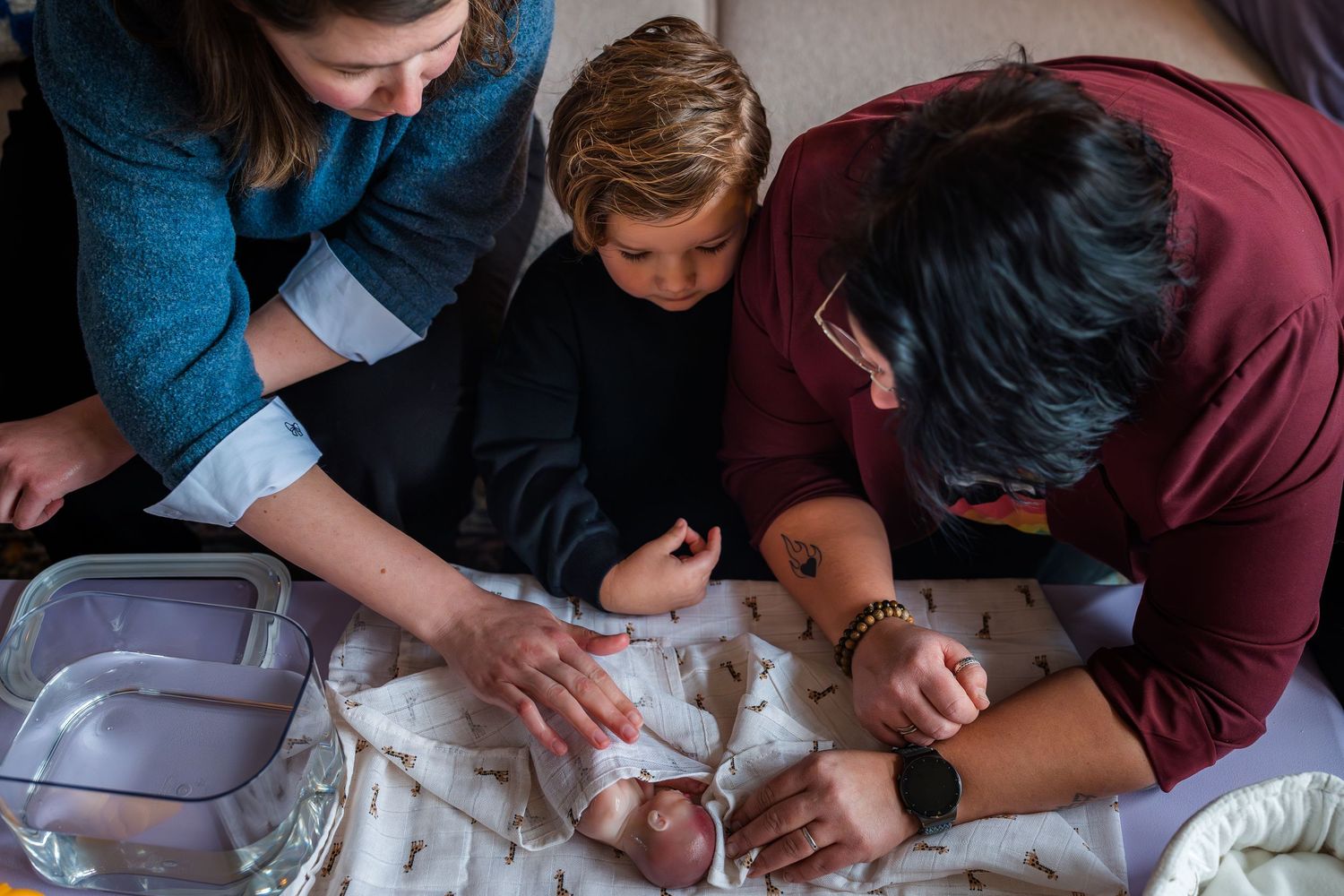 Family gathered around a newborn baby during a tender home birth moment, with a young child looking on curiously.