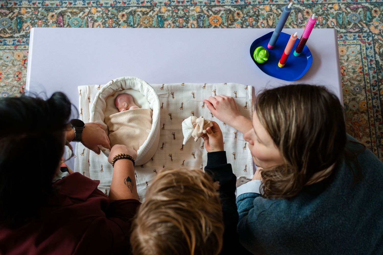 Overhead view of two adults and a child gathered around a newborn baby lying in a white bassinet on a table.