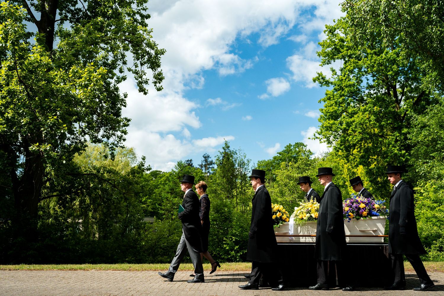 Pallbearers in black suits carry a flower-adorned casket along a tree-lined path during an outdoor funeral procession.