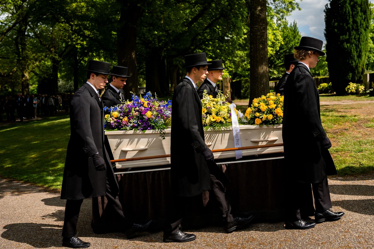 Pallbearers in black formal attire carry a white casket adorned with colorful flowers at an outdoor funeral.