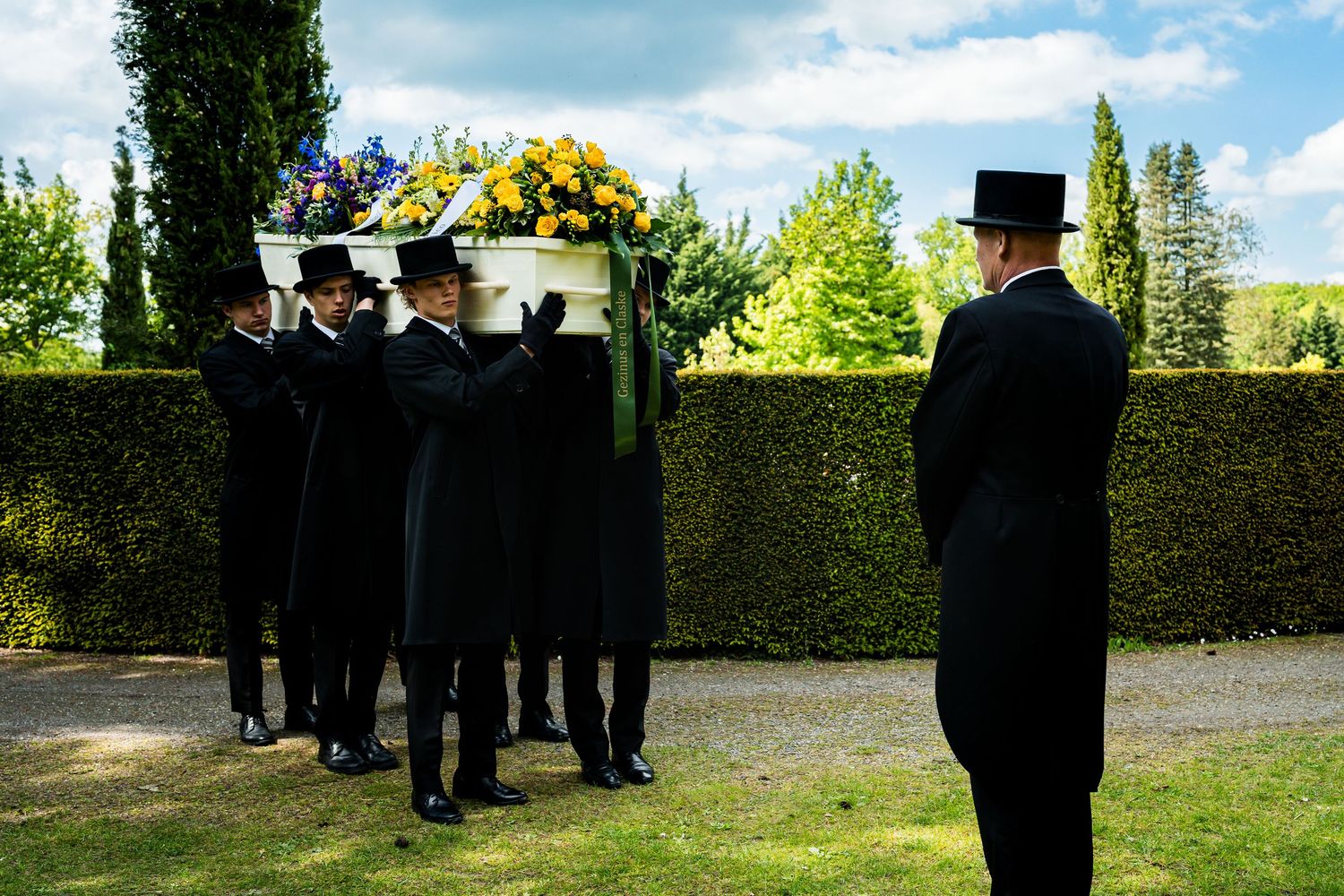 Pallbearers in black suits and top hats carry a white coffin with yellow flowers at an outdoor funeral ceremony.